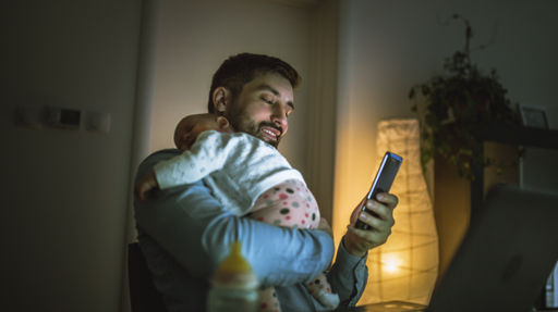 En man står upp med en liten bebis vilande på axeln. I sin lediga hand håller mannen en mobiltelefon.