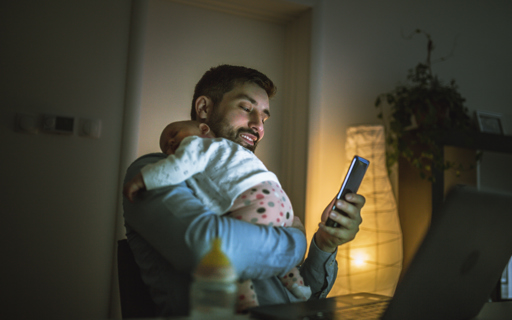 En man står upp med en liten bebis vilande på axeln. I sin lediga hand håller mannen en mobiltelefon.
