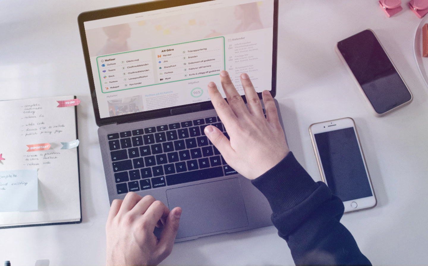 En person arbetar vid en bärbar dator på ett skrivbord. På skärmen visas ett digitalt kontrollpanel- eller intranätsgränssnitt med en att-göra-lista och olika systemikoner. På skrivbordet ligger även en mobiltelefon och anteckningar.