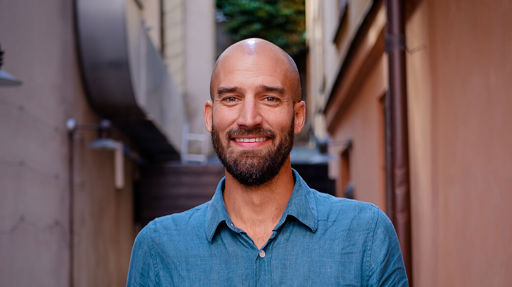 Kristoffer ler ståendes framför några äldre hus. Han har rakat huvud, mörkt skägg och en blå skjorta.