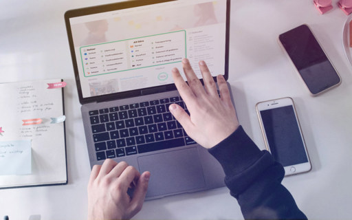 En person arbetar vid en bärbar dator på ett skrivbord. På skärmen visas ett digitalt kontrollpanel- eller intranätsgränssnitt med en att-göra-lista och olika systemikoner. På skrivbordet ligger även en mobiltelefon och anteckningar.